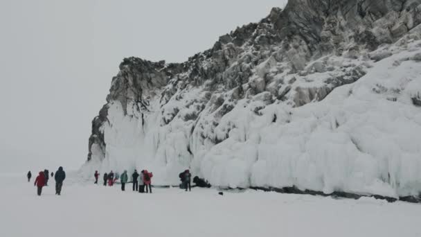 Baikal, Región de Irkutsk, Rusia - 18 de marzo de 2021: Un grupo de turistas viaja a lo largo del congelado lago Baikal y examina las cuevas y rocas cubiertas de carámbanos en la isla Olkhon. — Vídeo de stock