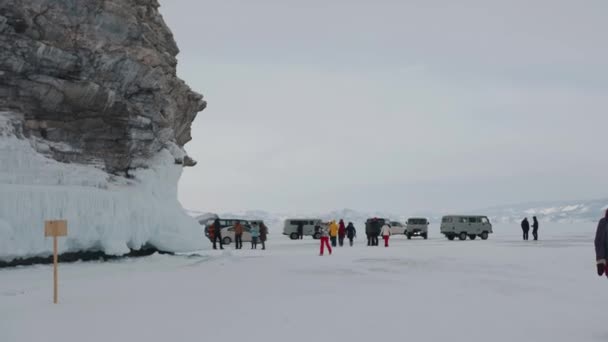 Bajkal, Irkutsk Region, Rusko - 18. března 2021: Mnoho turistů na břehu jezera Bajkal procházka a prohlédnout skály pokryté rampouchy a led a UAZ minibusy stojící v blízkosti — Stock video