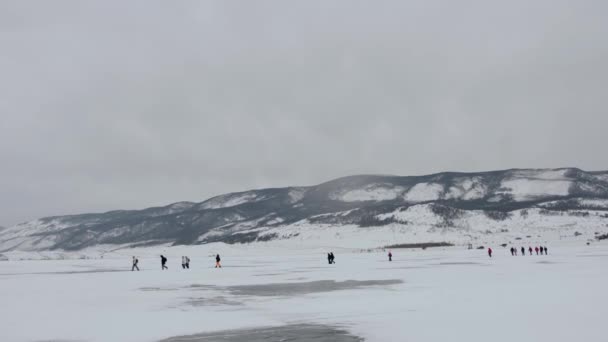 Een groep toeristen gaat de een na de ander op het sneeuw ijs van het Baikalmeer tegen de achtergrond van rotsen aan de kust — Stockvideo