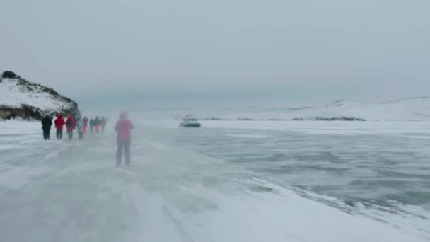 Bajkal, Irkutsk Region, Rusko - 18. března 2021: Skupina turistů kráčí podél břehu zamrzlého jezera Bajkal a dívá se na vznášedlo Khivus projíždějící kolem — Stock video