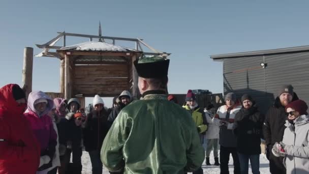 Région d'Irkoutsk, Russie - 18 mars 2021 : Un guide bouriate en costume national s'entretient avec des touristes dans un parc ethno sur la côte du Baïkal lors d'une excursion hivernale. Vue arrière — Video