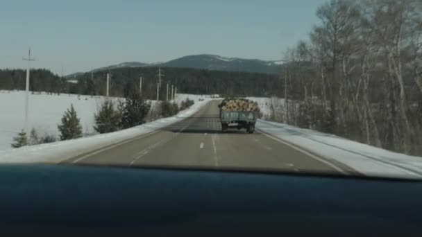 Vista attraverso il parabrezza di un'auto su un camion con tronchi sulla strada e un paesaggio invernale. Colpo palmare punto di vista della fotocamera — Video Stock