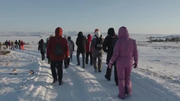 Irkoetsk regio, Rusland - 18 maart 2021: Een groep toeristen loopt langs een besneeuwde weg en bevroren Baikalmeer, omgeven door bergen aan de horizon. Achteraanzicht. Langzame beweging — Stockvideo
