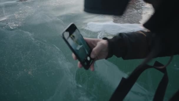 男はバイカル湖の透明氷の美しい質感の彼の携帯電話で写真を撮る。閉鎖 — ストック動画