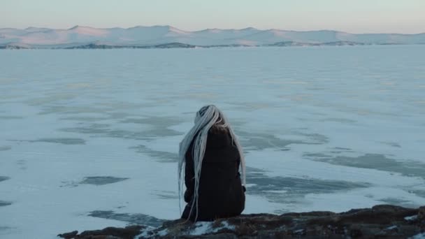 Mladá dívka sedí na skále a obdivuje nádhernou krajinu zamrzlého jezera Bajkal při západu slunce. Koncept svobody a klidu — Stock video