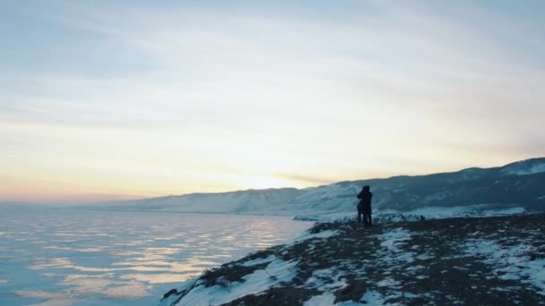 Eine Gruppe Touristen steht auf einem Hügel und genießt den Blick auf die Bergküste des zugefrorenen Baikalsees bei Sonnenuntergang — Stockvideo