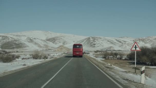 Baikal, Región de Irkutsk, Rusia - 18 de marzo de 2021: Vista a través del parabrisas de un automóvil en un autobús rojo en la carretera y las montañas cubiertas de nieve. pov de mano de la cámara — Vídeo de stock