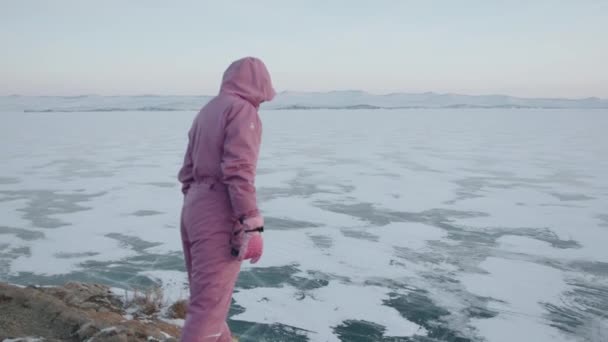 Іркутська область (Росія) 18 березня 2021 р.: молода дівчина в рожевому покритті і сонячні окуляри стоять на краю скелі і схиляється над тим, щоб дивитися на замерзле озеро Байкал. — стокове відео