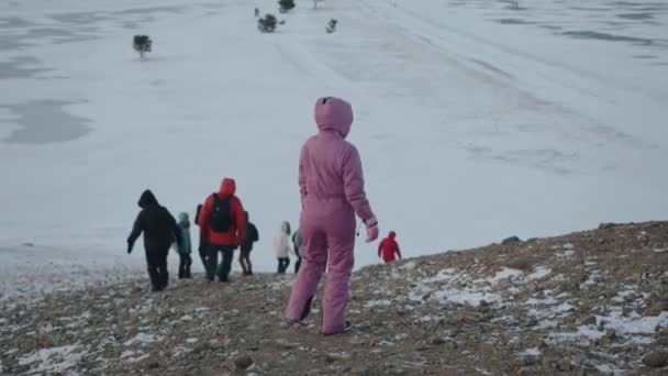 Región de Irkutsk, Rusia - 18 de marzo de 2021: Un grupo de turistas desciende la colina y va al lago cubierto de hielo Baikal, rodeado de montañas. Vista trasera — Vídeos de Stock