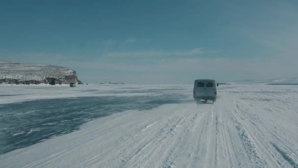 Coluna de microônibus UAZ em passeio de alta velocidade no gelo transparente do Lago Baikal passado as montanhas rochosas na costa — Vídeo de Stock