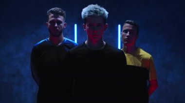 Üç çekici genç futbolcu karanlık bir stüdyoda neon ışıkların arka planına karşı dururlar ve içlerinden biri topu elleriyle yakalar. 4K