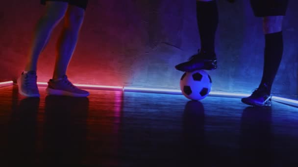 Deux joueurs de football donnent un coup de pied au ballon dans un studio sombre avec éclairage au néon. Gros plan des pieds des athlètes. 4K — Video