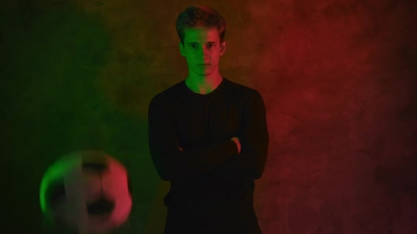 Ένας όμορφος νεαρός ποδοσφαιριστής με μια μαύρη φανέλα στέκεται με τα χέρια του διπλωμένα στο στήθος του στο φως του νέον και μια μπάλα πηδάει μπροστά του. 4K — Αρχείο Βίντεο
