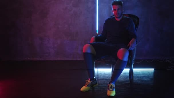 Un guapo jugador de fútbol barbudo se sienta en una silla en un estudio oscuro con iluminación de neón y detiene una bola rodante con su pie. 4K — Vídeos de Stock