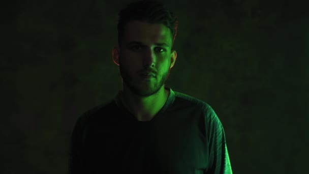 Ett porträtt av en brutal skäggig ung idrottsman i ett mörkt rum i neonljus med händerna vikta på bröstet. 4K — Stockvideo