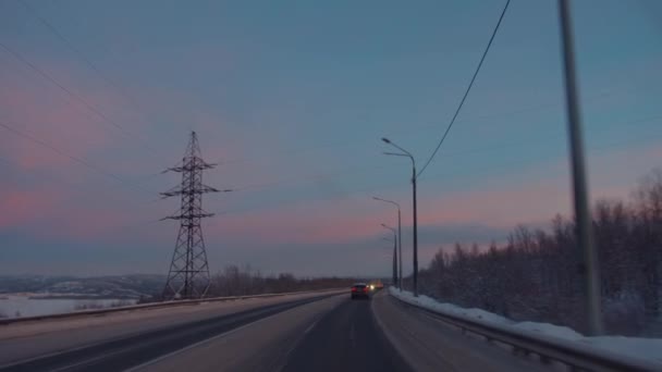 Vista através do pára-brisas do movimento de carros em uma estrada suburbana de inverno e os pilões da linha de energia ao longo da estrada ao pôr do sol — Vídeo de Stock