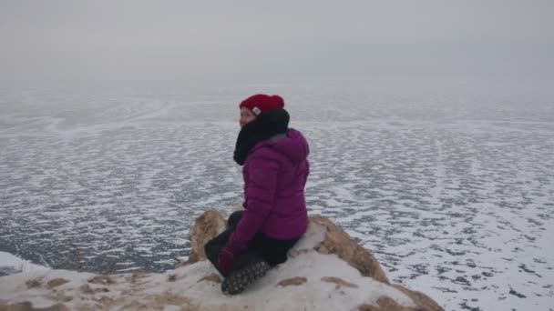 Mladá dívka sedí na skále na pozadí obrovských rozloh zamrzlého jezera Bajkal v oparu — Stock video