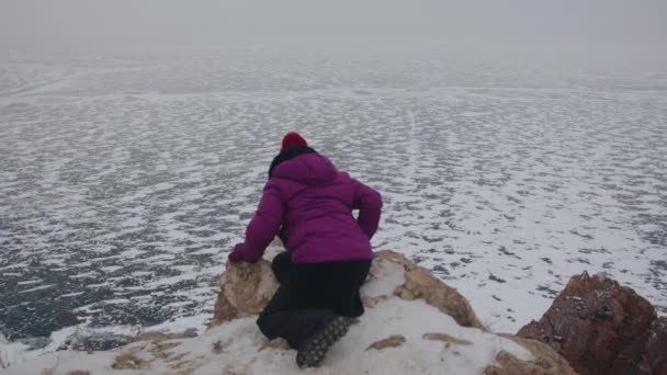氷で覆われたバイカル湖の海岸を見下ろすと、若い女性が高い岩の上に座って曲がります — ストック動画