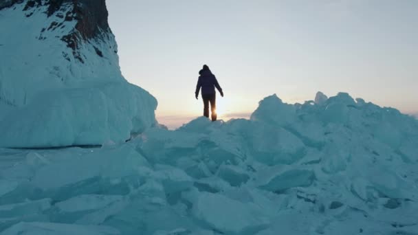 Una giovane ragazza cammina lungo i blocchi di ghiaccio blu e bianco del lago Baikal accanto alla roccia sullo sfondo di un tramonto. Rallentatore — Video Stock