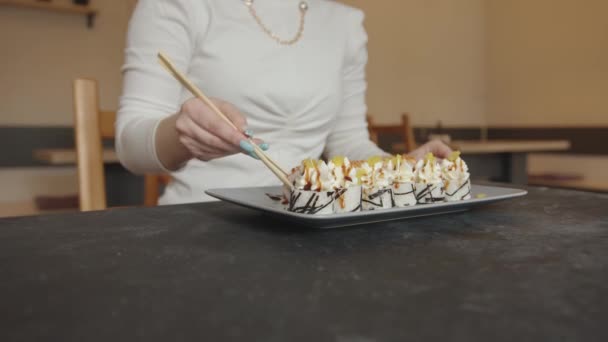 Uma jovem espetacular senta-se em um café e leva sushi maki de um prato e demonstra-o para a câmera. Movimento lento — Vídeo de Stock