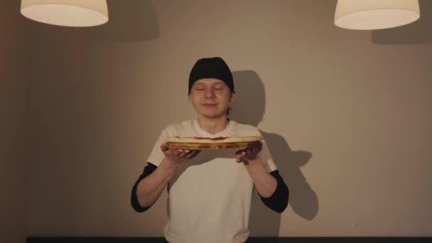 Un joven chef en un pañuelo sostiene una pizza en un café y la huele disfrutando del aroma — Vídeos de Stock