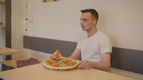 Joven hombre atractivo en una camiseta blanca se sienta en una mesa en un café y come pizza — Vídeo de stock