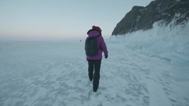 Una joven camina a lo largo del congelado lago Baikal pasando por bloques de hielo y montañas rocosas en la orilla. Vista trasera. Movimiento lento — Vídeos de Stock