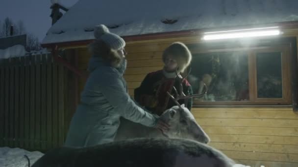 Murmańsk, Rosja - 10 stycznia 2021 r.: Młoda dziewczyna słucha opowieści o kobiecie-przewodniku w ubraniach narodowych i głaszcze jelenia na farmie w zimowy wieczór — Wideo stockowe