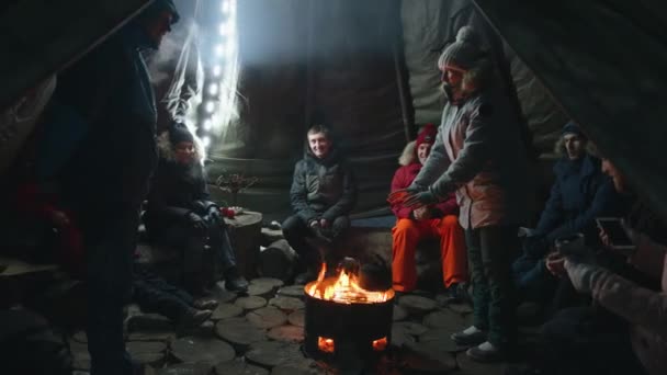 러시아, 무르만스크 - 2021 년 1 월 10 일: 한 무리의 관광객들 이 겨울 여행중 텐트 안의 넓은 난로에 앉아 불옆에서 몸을 따뜻하게 한다 — 비디오