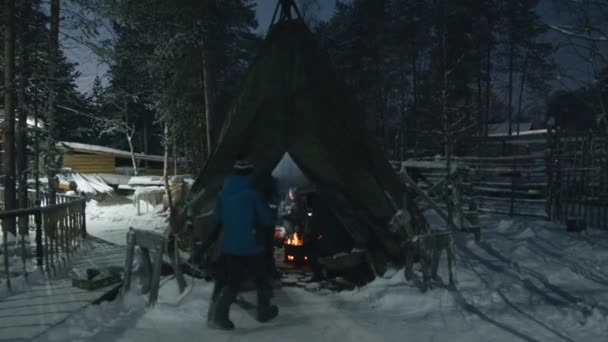 Um jovem entra na tenda wigwam em um pátio rural, onde um grupo de turistas se senta ao redor da lareira com um fogo em uma noite de inverno. — Vídeo de Stock