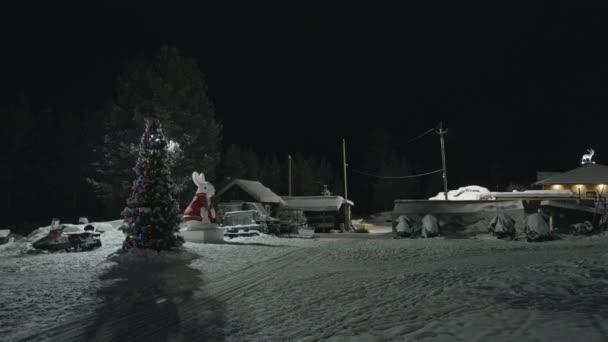 Двор деревни в зимний вечер украшен елой в красочных гирляндах и большой фигурой рождественского кролика — стоковое видео