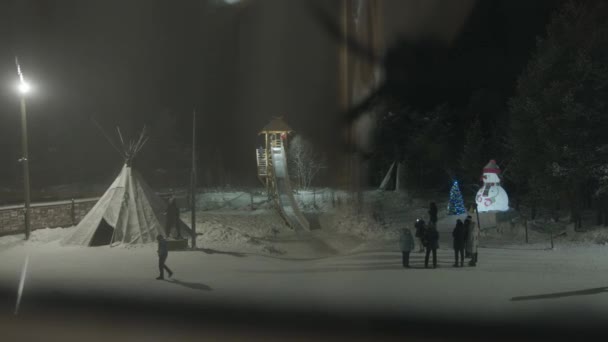 Группа туристов осматривает детскую площадку, украшенную на Рождество, и катается с деревянной горки зимним вечером — стоковое видео