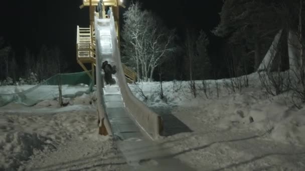 Murmansk, Rusko - 10. ledna 2021: Mladí muži se poflakují a jezdí na zádech z velkého dřevěného skluzavky za zimního večera — Stock video