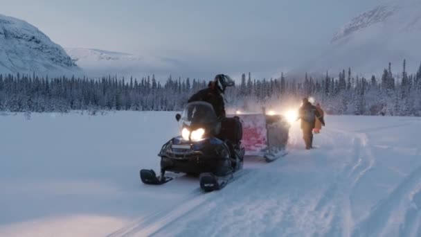 Murmansk, Rússia - 10 de janeiro de 2021: Um jovem em um capacete monta uma moto de neve com um trenó de madeira preso para transportar turistas e acena sua mão. Movimento lento — Vídeo de Stock