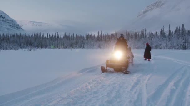 Murmansk, Ρωσία - 10 Ιανουαρίου 2021: Ένας νεαρός άνδρας με κράνος οδηγεί ένα snowmobile με ένα ξύλινο έλκηθρο που συνδέεται με τη μεταφορά τουριστών και κύματα το χέρι του. Αργή κίνηση — Αρχείο Βίντεο