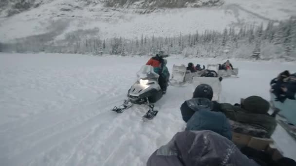 Murmansk region, Russia - January 10, 2021: A man driving a snowmobile carries tourists in a wooden sleigh through a snowy field — стоковое видео