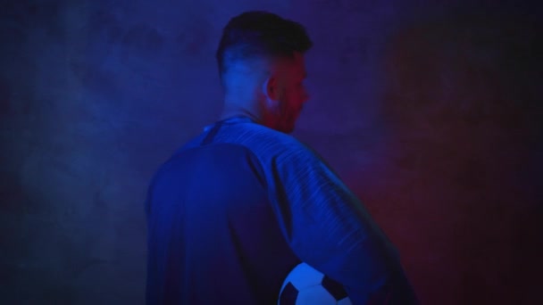 Stilig skäggig ung fotbollsspelare står med ryggen i en mörk studio i neonljus med en boll i handen och vänder blicken intensivt mot kameran. 4K — Stockvideo