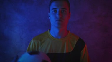 Çekici genç bir futbolcu karanlık bir stüdyoda neon ışığında duruyor ve bir elden diğerine top atıyor. 4K