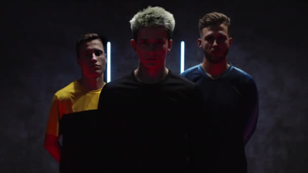 Jonge voetballers staan tegen de achtergrond van een muur met strepen van neon lampen in een donkere studio en een van hen houdt een voetbal in de hand. 4K — Stockvideo