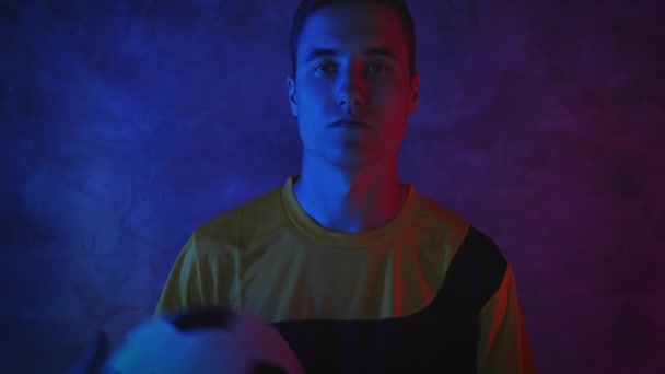 Un joven jugador de fútbol atractivo se encuentra en un estudio oscuro con luz de neón y lanza una pelota de una mano a otra. 4K — Vídeos de Stock