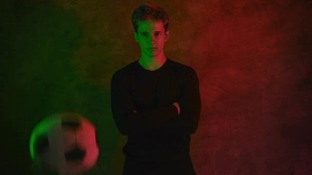 A handsome young soccer player in a black jersey stands with his arms folded across his chest in the neon light and a ball jumps in front of him. 4K — Stock Video