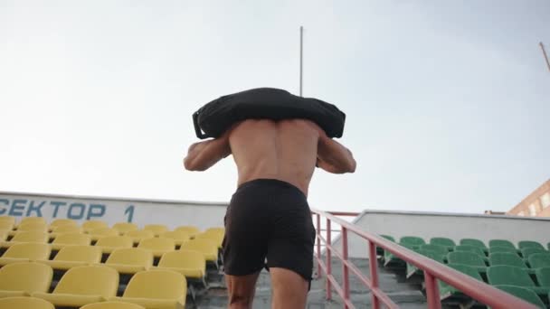 Un bodybuilder in pantaloncini neri corre sui gradini dello stadio cittadino con un sacco di sabbia sulle spalle. Al rallentatore. A seguito di colpo — Video Stock