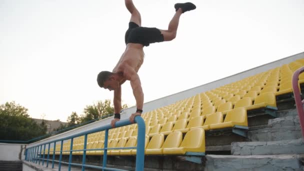 En muskulös man går in för sport i arenan och gör en handställning lutar sig på räcket i stället. Långsamma rörelser — Stockvideo