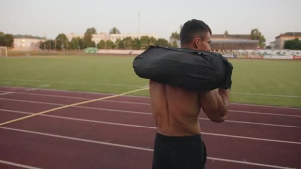Um homem musculoso treinando no estádio e caminha com um saco de areia em seus ombros na esteira. Movimento lento. Seguinte tiro — Vídeo de Stock
