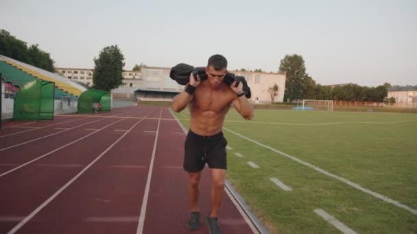 Stadyumda çıplak vücutlu kaslı bir adam antrenman yapıyor ve omuzlarında kum torbasıyla koşu bandında koşuyor. Yavaş çekim — Stok video