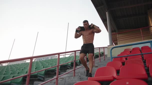 En muskulös man i svarta shorts tränar på stadion och springer ner för trappan till läktaren med en sandsäck på axlarna. Långsamma rörelser — Stockvideo
