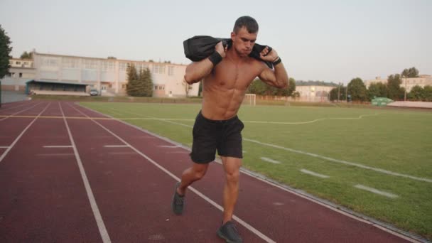 Fisiculturista em shorts pretos treina no estádio e lunges com um saco de areia em seus ombros — Vídeo de Stock