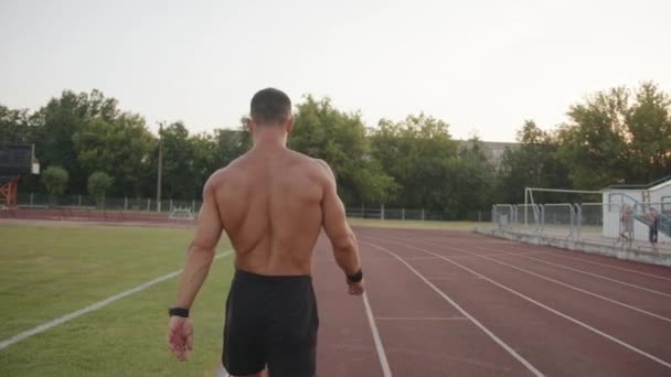 En muskulös man med en naken överkropp går längs löpbandet i stadens stadion. Bakåt. Långsamma rörelser — Stockvideo