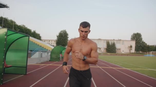 Un culturista torso desnudo corre a lo largo de la cinta de correr del estadio de la ciudad con una expresión enfocada en su cara. Movimiento lento — Vídeos de Stock