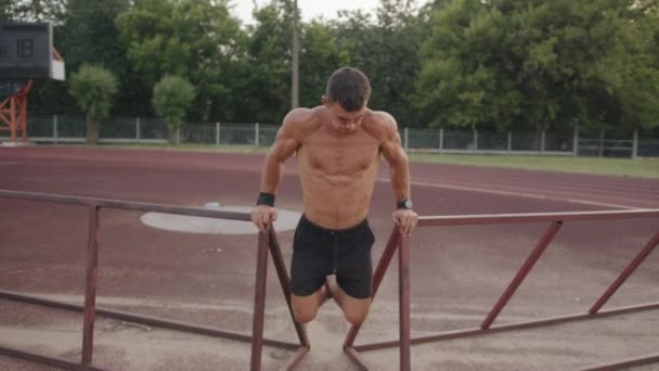 Hombre musculoso en pantalones cortos negros entrenando en el estadio de la ciudad y haciendo flexiones en bastidores metálicos. Concepto de estilo activo — Vídeo de stock